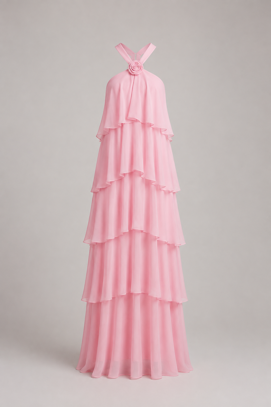 A light pink halter-neck maxi dress with a tiered ruffle design and a floral embellishment at the neckline, shown on a neutral gray background.