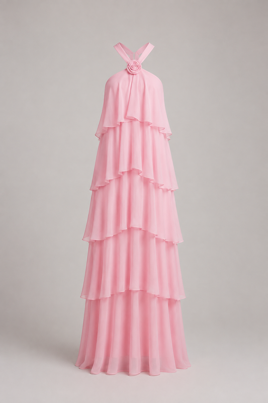 A light pink halter-neck maxi dress with a tiered ruffle design and a floral embellishment at the neckline, shown on a neutral gray background.